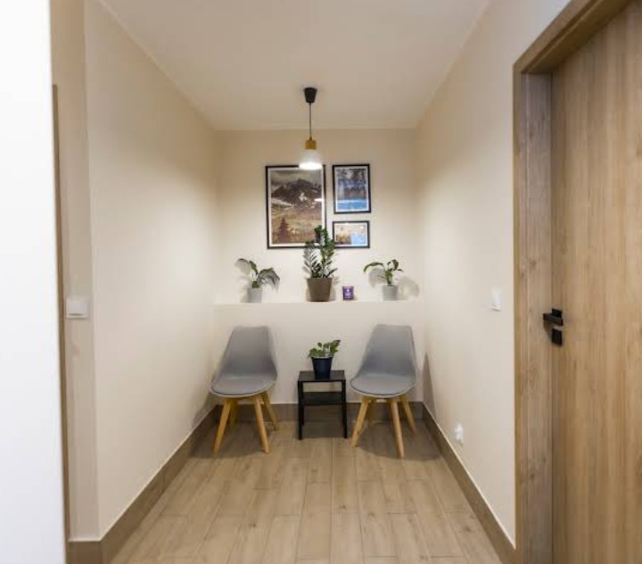 Jasne wnętrze poczekalni z dwoma szarymi krzesłami, stolikiem, roślinami doniczkowymi i obrazami na ścianie, oświetlone wiszącą lampą z żółtym kloszem.