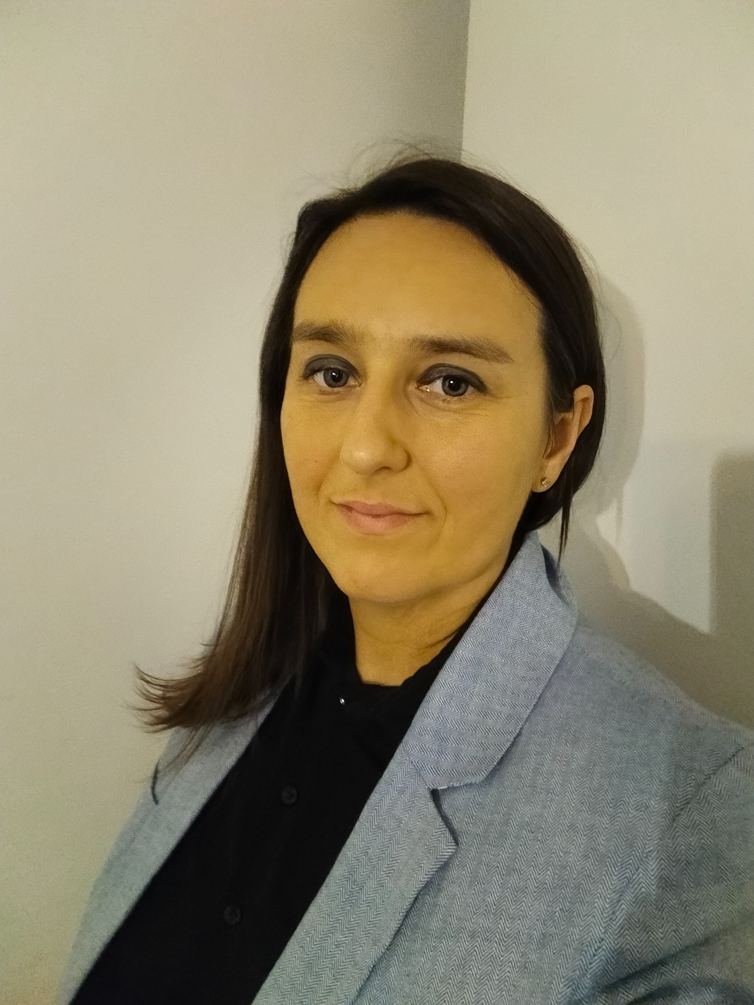 Portret kobiety z ciemnymi włosami, uśmiechniętej, ubranej w czarną koszulę i szary żakiet w jodełkę, stojącej na tle jasnej ściany.