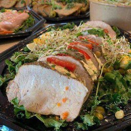 Pełna Micha - Plastry pieczonego mięsa na czarnej tacy, udekorowane zielonymi liśćmi, kiełkami i pomidorami, z sałatką w tle.