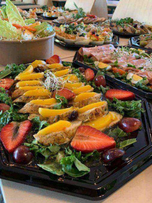 Różnorodne dania w czarnych pojemnikach cateringowych: sałatka z kurczakiem i mango, sałatka z szynką i jajkiem, sałatki warzywne w papierowych misach, całość na stole.