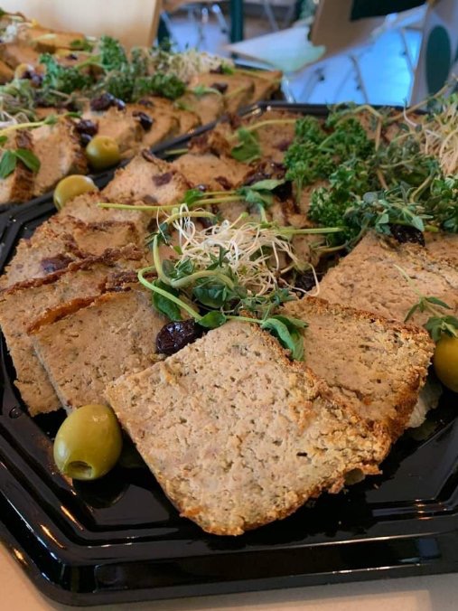 Półmisek z pasztetem pokrojonym w plastry, udekorowany oliwkami, świeżymi ziołami i kiełkami, na czarnej, lakierowanej tacy cateringowej.