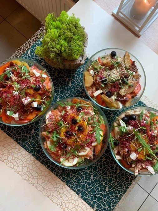 Cztery szklane miski z kolorowymi sałatkami na dekoracyjnym obrusie, z mchem w doniczce i świecą w tle.