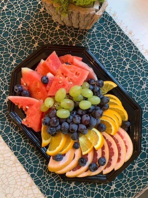 Czarny, ośmiokątny talerz z aranżacją owoców: arbuza, zielonych i fioletowych winogron, pomarańczy, grejpfruta i borówek, ułożonych na zielonym obrusie z białym wzorem, obok dekoracja z mchu.