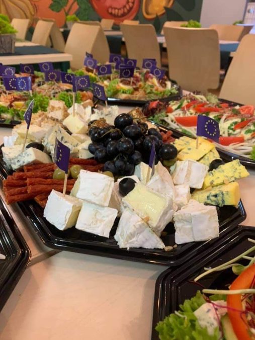 Ułożone na czarnych tackach sery pleśniowe, brie i winogrona ozdobione flagami UE, na stole podczas wydarzenia.