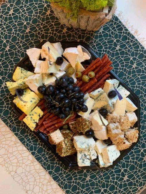 Dekoracyjna czarna taca z selekcją serów, winogron, oliwek i cienkich kabanosów ułożona na zielonej serwecie o wzorze pajęczyny, z doniczką z mchem w tle.