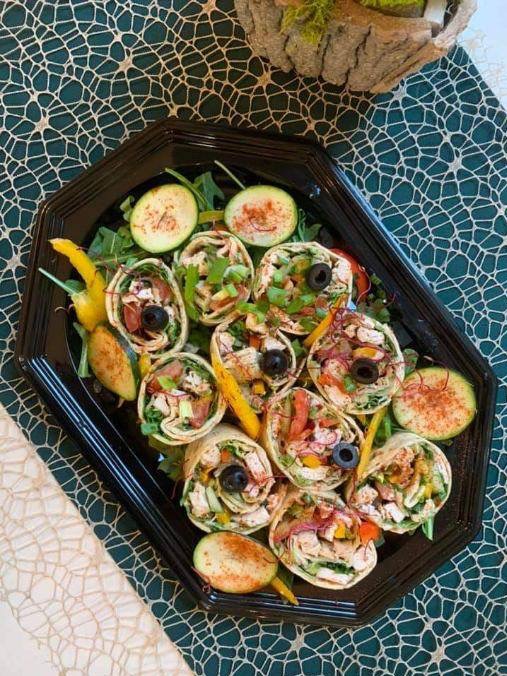 Czarna taca z roladkami tortilli, z kurczakiem, warzywami i oliwkami, udekorowana plasterkami ogórka posypanymi przyprawą, na tle wzorzystej serwety w odcieniach zieleni.