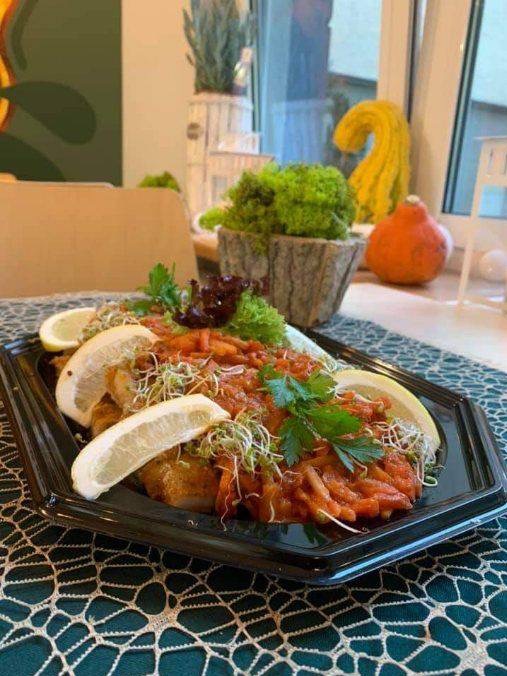 Elegancko podane danie rybne z sosem pomidorowym, kiełkami i cytryną na czarnym talerzu, Warszawa. W tle dekoracje okienne z dyniami i roślinami.