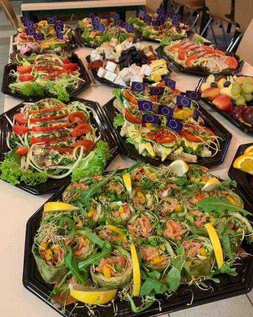 Różnorodne przekąski na czarnych tacach: rolki z łososiem i cytryną, sałatki warzywne z pomidorami, sery z owocami, dekorowane flagami UE.