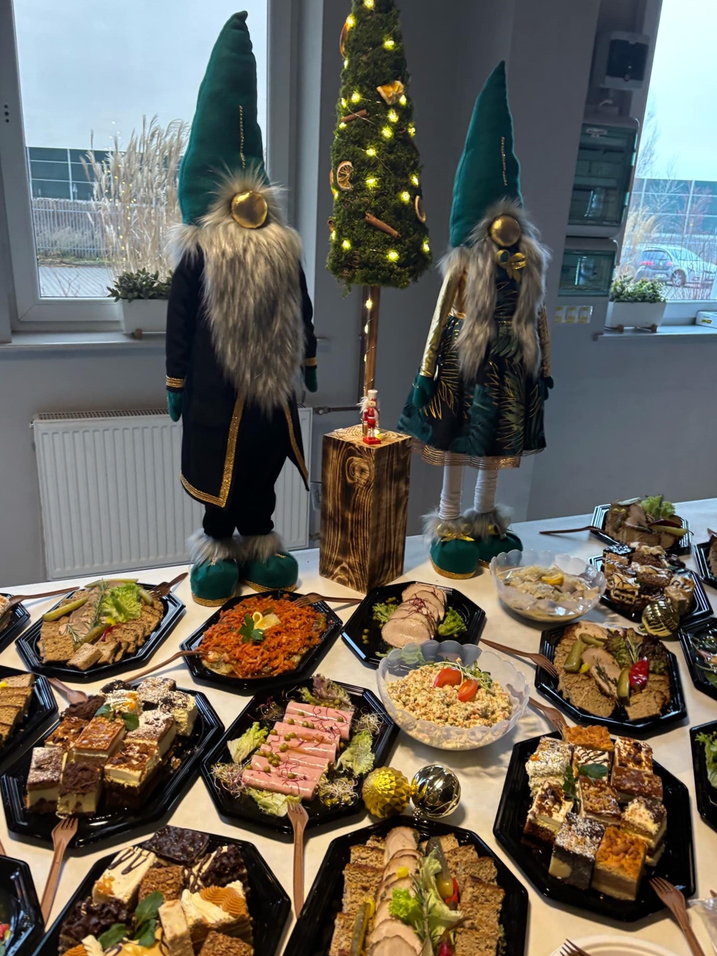 Stół z różnorodnymi potrawami, dekoracjami świątecznymi w postaci dwóch gnomów w zielonych czapkach i małej choinki, ustawiony w jasnym wnętrzu.