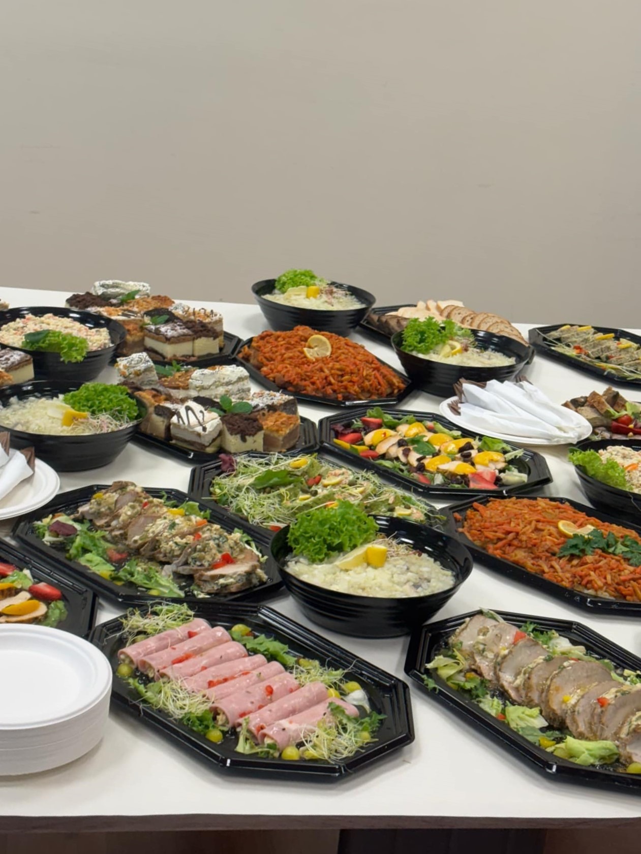 Stół z różnorodnymi potrawami w czarnych naczyniach: sałatki z owocami, rolady mięsne, torty i dania z makaronem udekorowane zieleniną i cytryną.