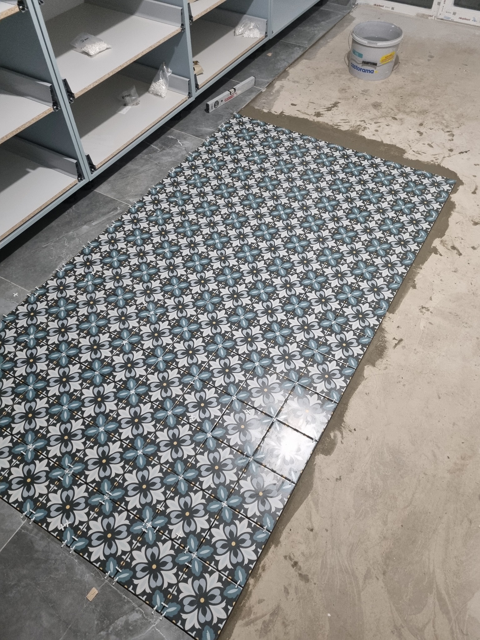 Układanie wzorzystych płytek podłogowych w trakcie remontu, widoczny poziomica, krzyżyki dystansowe i wiadro z zaprawą.