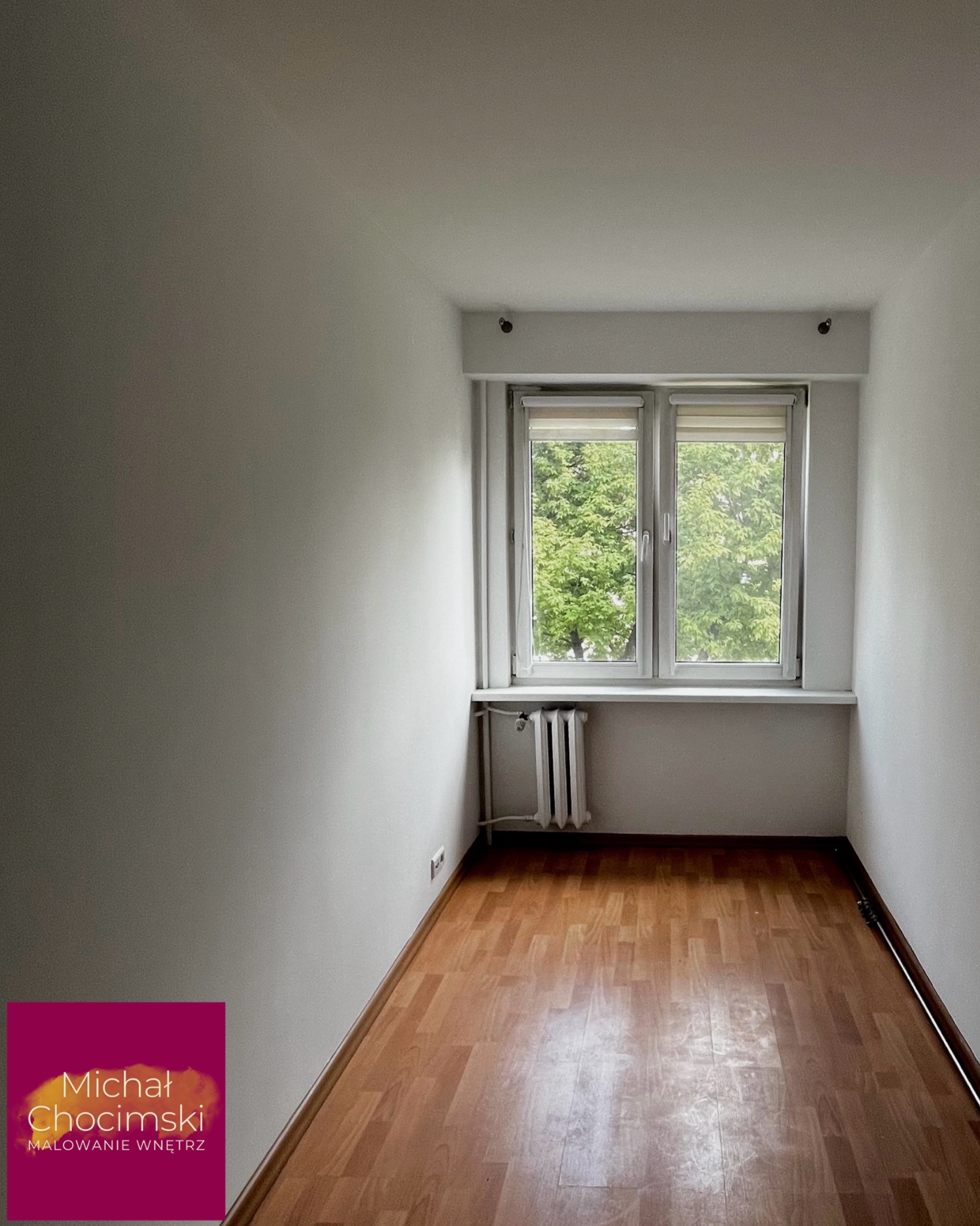 Pusty pokój z oknem, świeżo po odnowieniu. Białe ściany, drewniana podłoga. Widok na zieleń za oknem. Minimalistyczne wnętrze po remoncie.