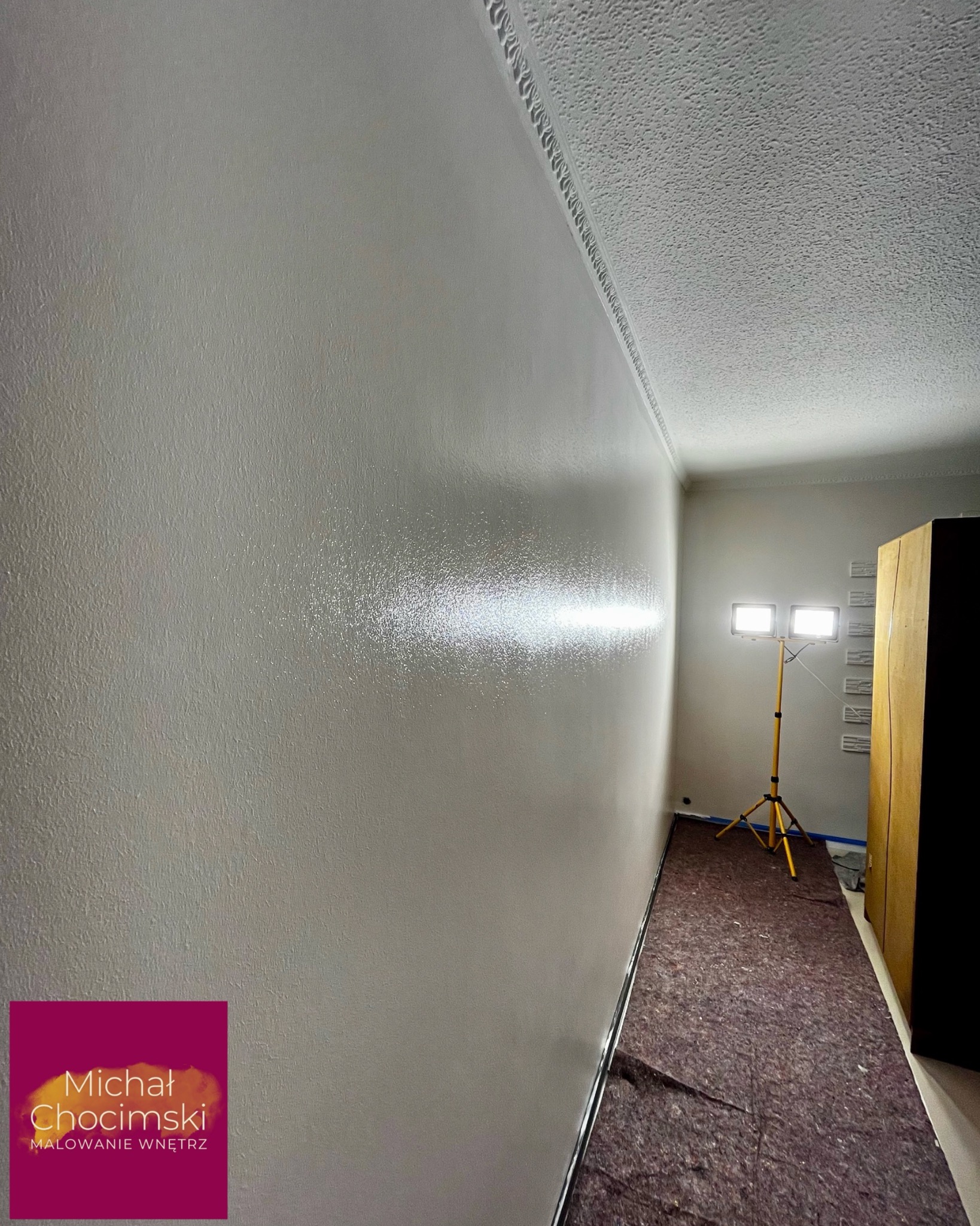 Świeżo pomalowana ściana w jasnym odcieniu z dekoracyjną listwą sufitową. Na podłodze zabezpieczenie. Widoczne oświetlenie robocze.