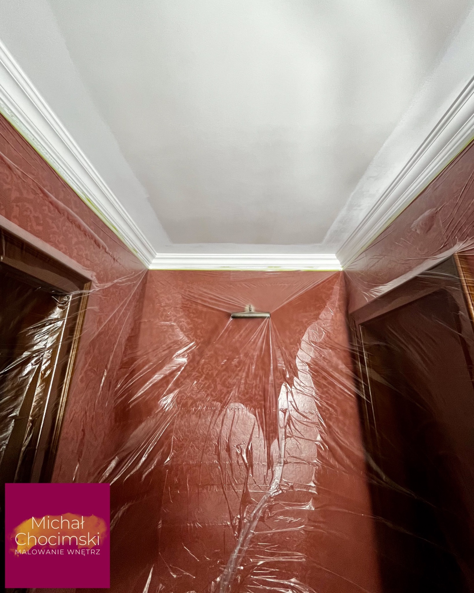 Malowanie sufitu z dekoracyjną sztukaterią, zabezpieczone ściany folią malarską. Widoczne fragmenty drzwi i logo firmy malarskiej w rogu zdjęcia.