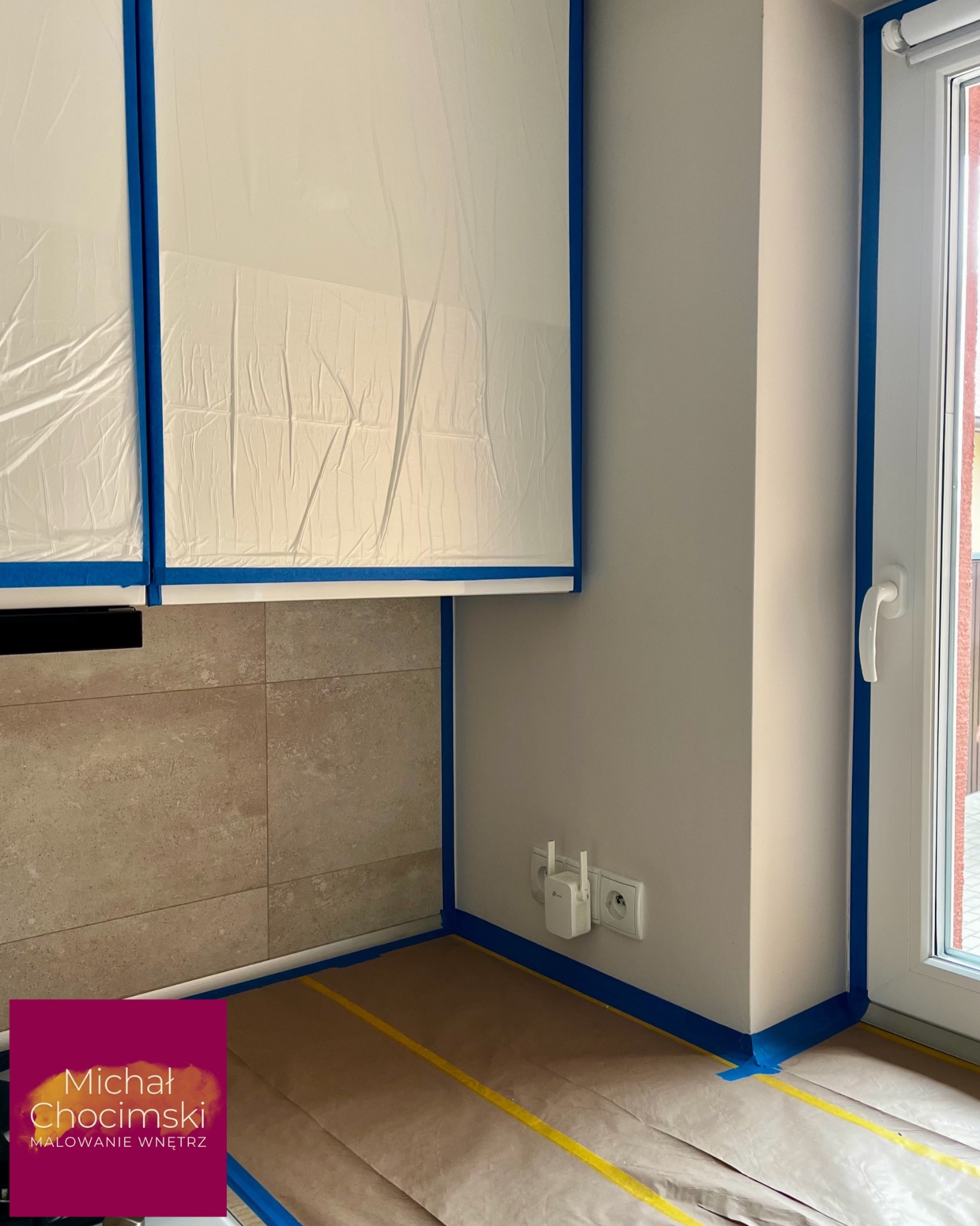 Przygotowania do malowania: zabezpieczone meble folią, podłoga kartonem i taśmą malarską. Gniazdka i listwy przypodłogowe oklejone taśmą. Widok na okno i ścianę.