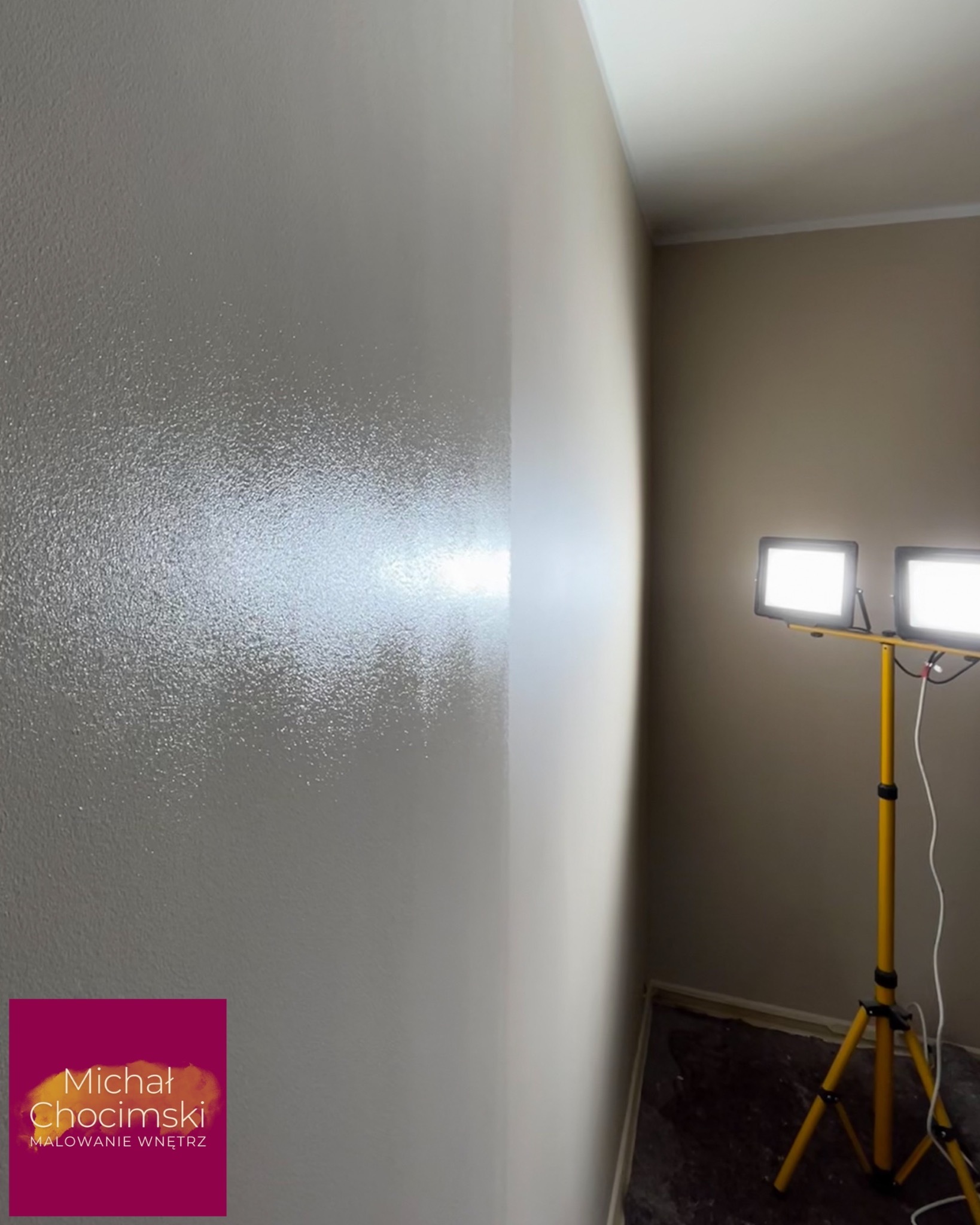 Świeżo pomalowana ściana w odcieniach szarości, oświetlona lampą budowlaną. Widoczna faktura farby, gładka powierzchnia. Efekt malowania wnętrza.