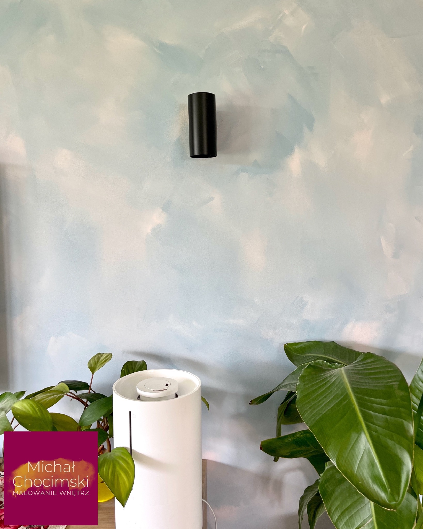 Ściana po malowaniu z efektem chmur, oświetlona czarnym kinkietem. W tle widoczne rośliny doniczkowe i nawilżacz powietrza. Subtelne przejścia kolorów i faktura.