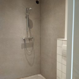 Lucas - Minimalistyczna kabina prysznicowa z szarymi, matowymi płytkami wielkoformatowymi, odpływem liniowym w rogu i białą toaletą podwieszaną z ukrytą spłuczką.