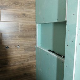 Lucas - Ściana z płyty gipsowo-kartonowej z wnęką, widoczne otwory na wkręty, obok ściana z brązowych płytek imitujących drewno z przygotowanymi otworami na armaturę