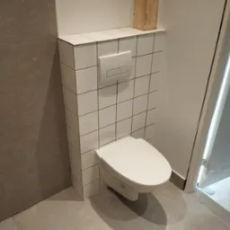 Ściana z białych płytek kwadratowych z ukrytym stelażem WC i przyciskiem spłukującym, szare płytki podłogowe, widoczny fragment drzwi.