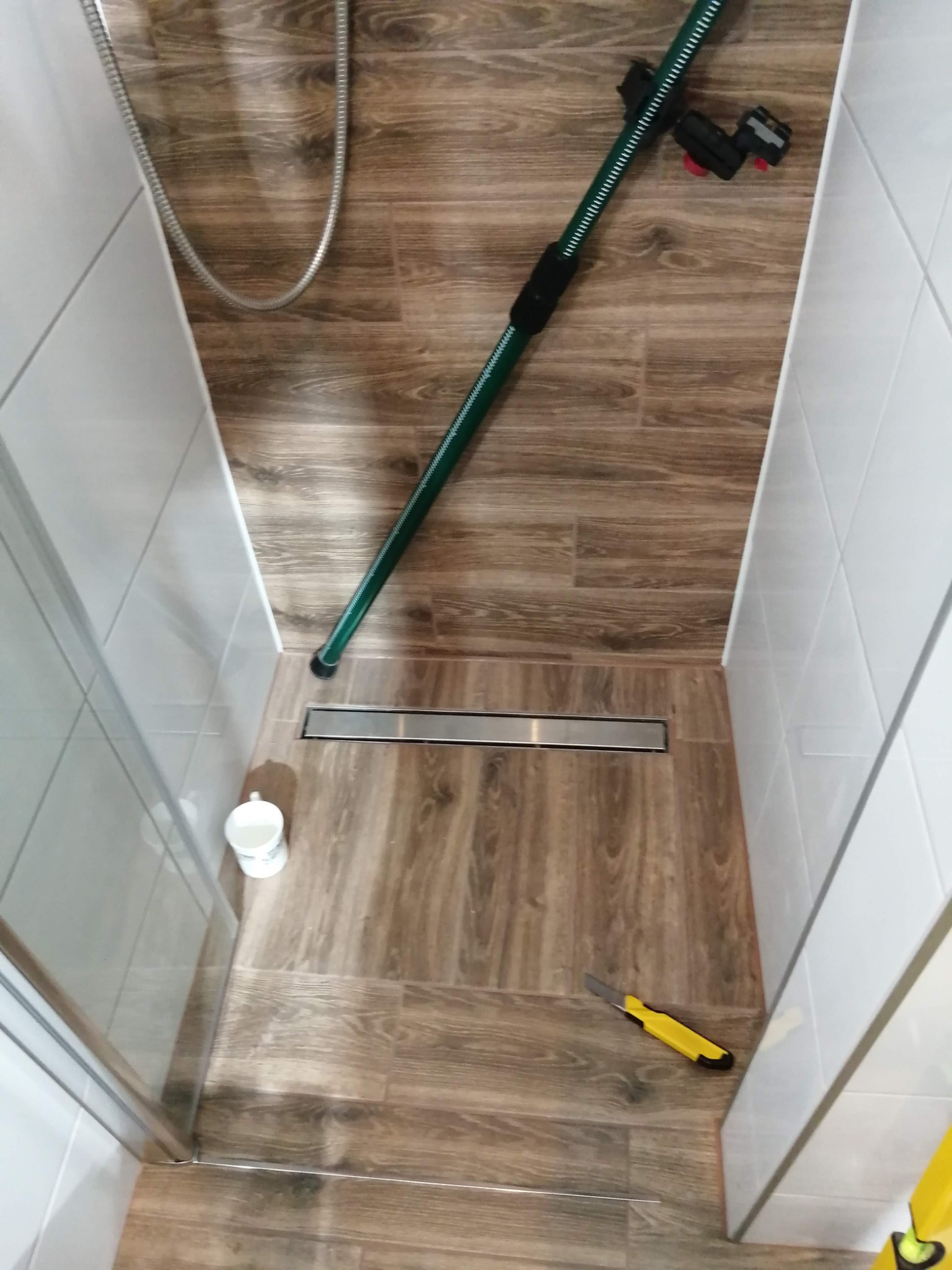 Ukończony prysznic typu walk-in z odpływem liniowym, wykończony płytkami imitującymi drewno na podłodze i ścianie, widoczny wężyk poziomujący, kubek z pozostałościami zaprawy i nóż do cięcia płytek.