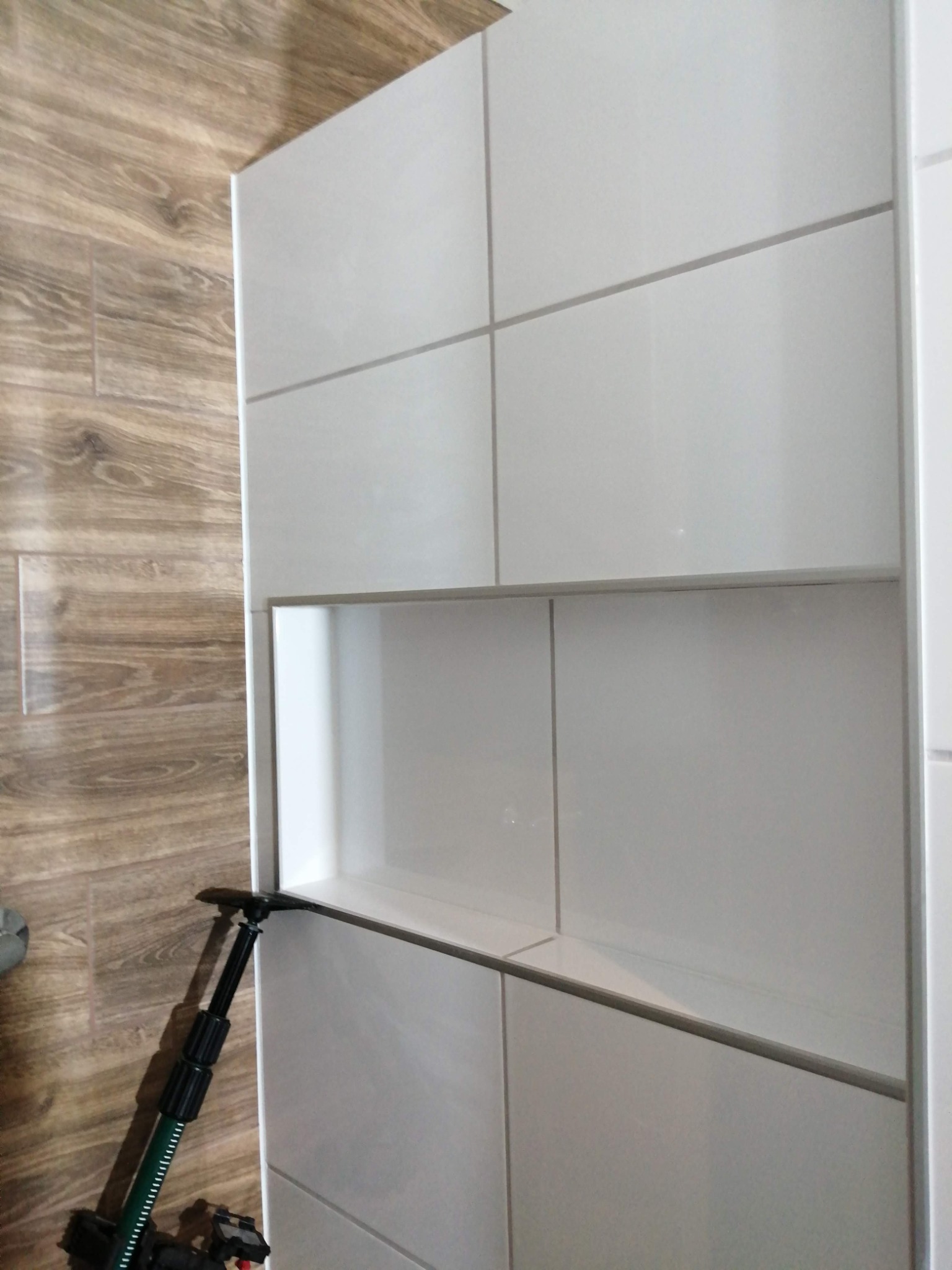 Biała, błyszcząca ściana z płytek w łazience, z wnęką wykończoną tymi samymi płytkami, widoczny fragment podłogi z płytek imitujących drewno oraz narzędzie do układania płytek.