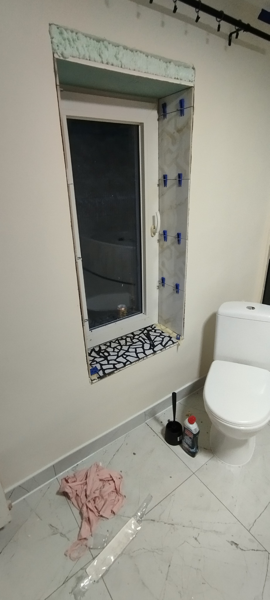 Montaż płytek mozaikowych na wnęce okiennej w łazience, widoczne niebieskie klipsy poziomujące, pianka montażowa wokół okna, biała toaleta, szczotka do WC i chemia budowlana na marmurowej podłodze.