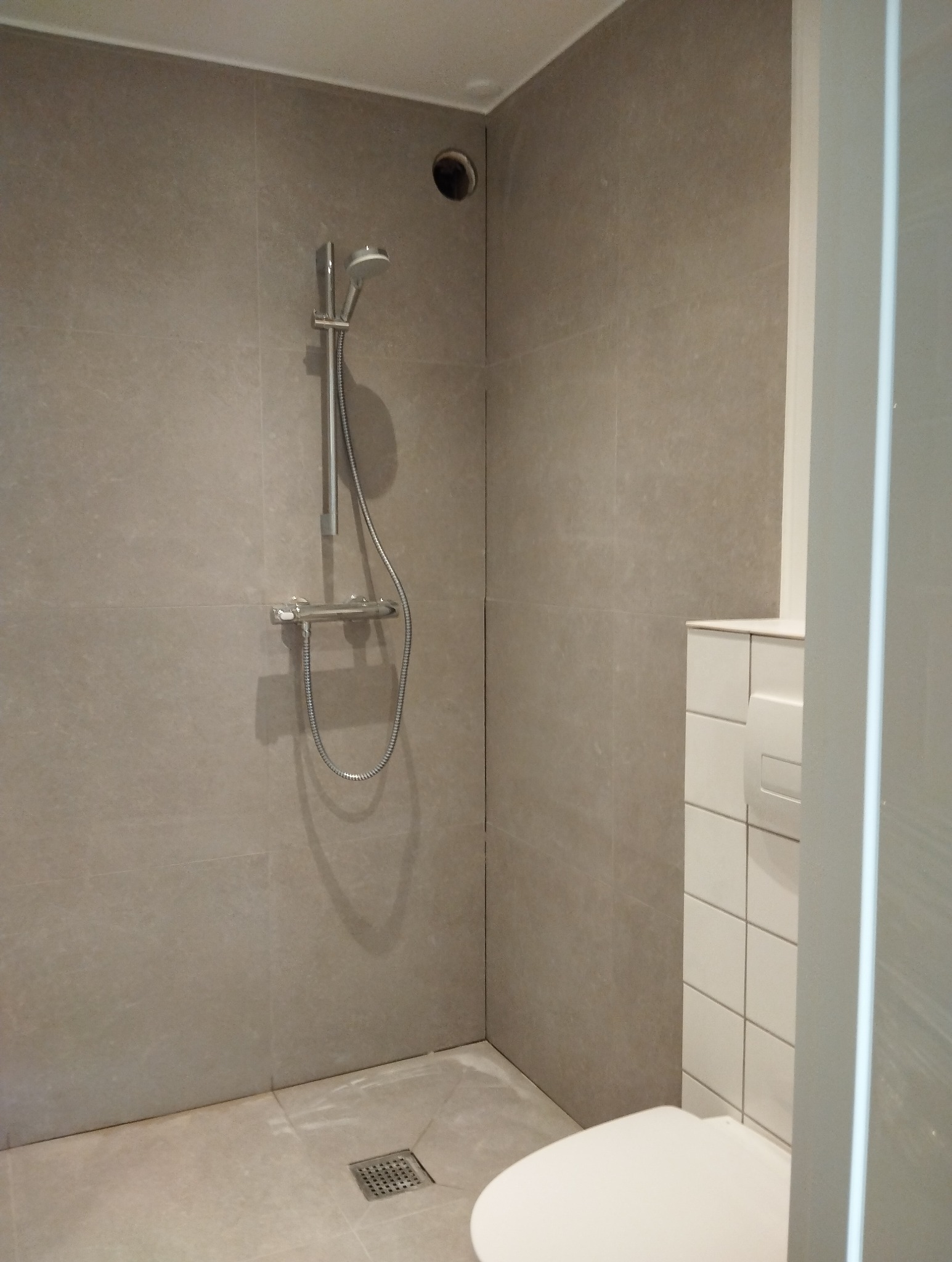 Minimalistyczna kabina prysznicowa z szarymi, matowymi płytkami wielkoformatowymi, odpływem liniowym w rogu i białą toaletą podwieszaną z ukrytą spłuczką.