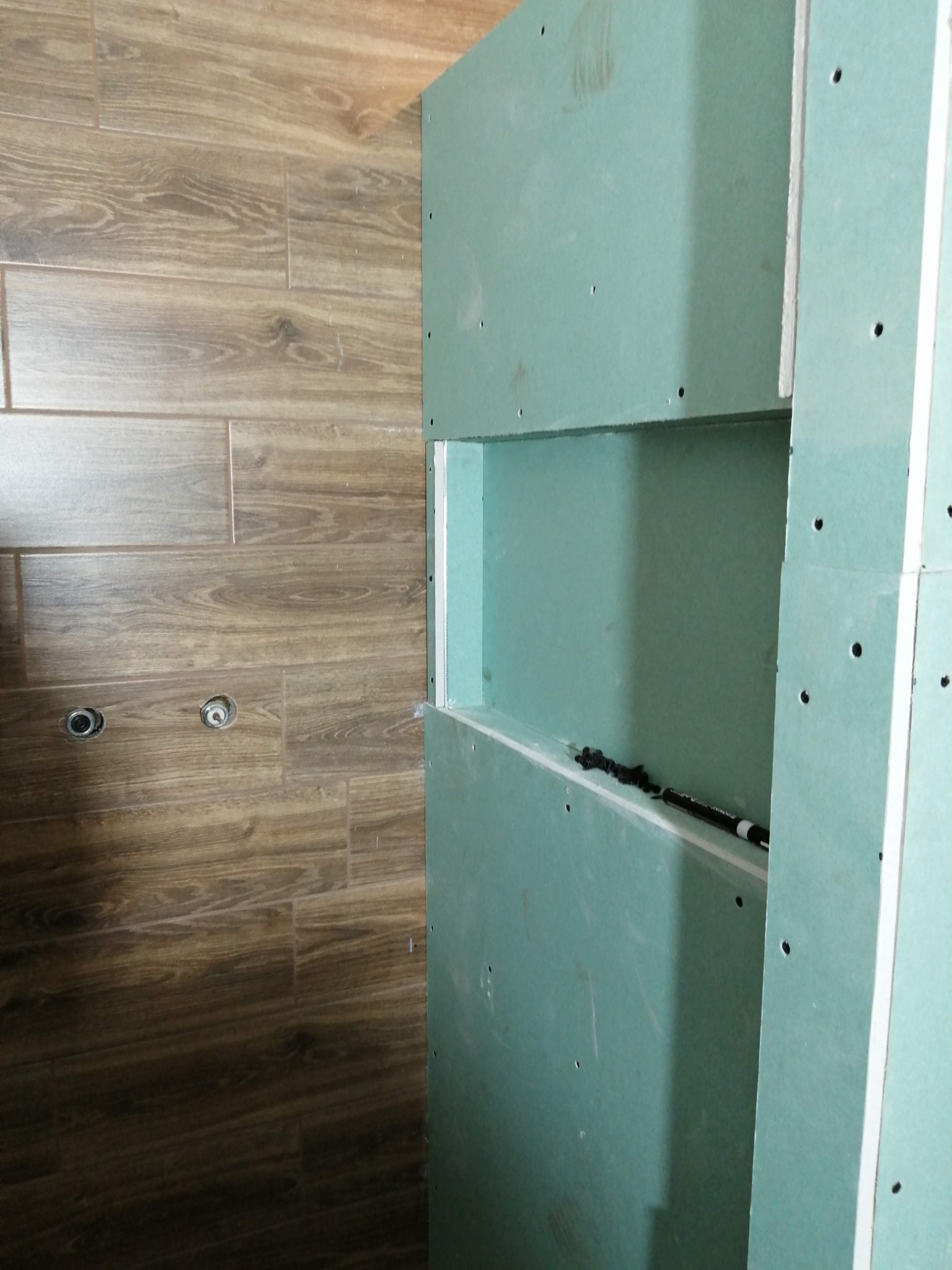 Ściana z płyty gipsowo-kartonowej z wnęką, widoczne otwory na wkręty, obok ściana z brązowych płytek imitujących drewno z przygotowanymi otworami na armaturę