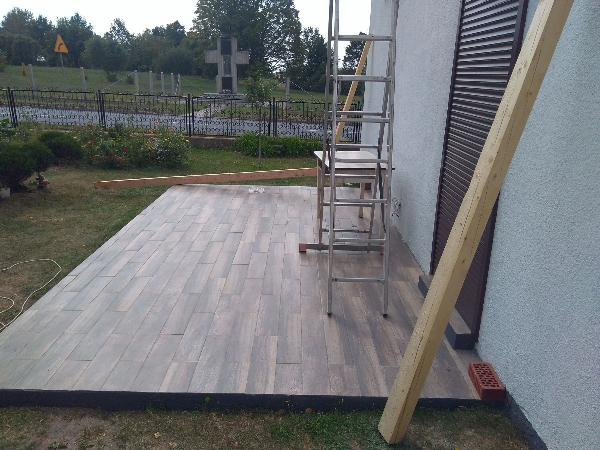 Wyłożony płytkami taras zewnętrzny z widocznymi deskami do montażu, drabina oparta o ścianę budynku, widok na ogród z pomnikiem w tle.