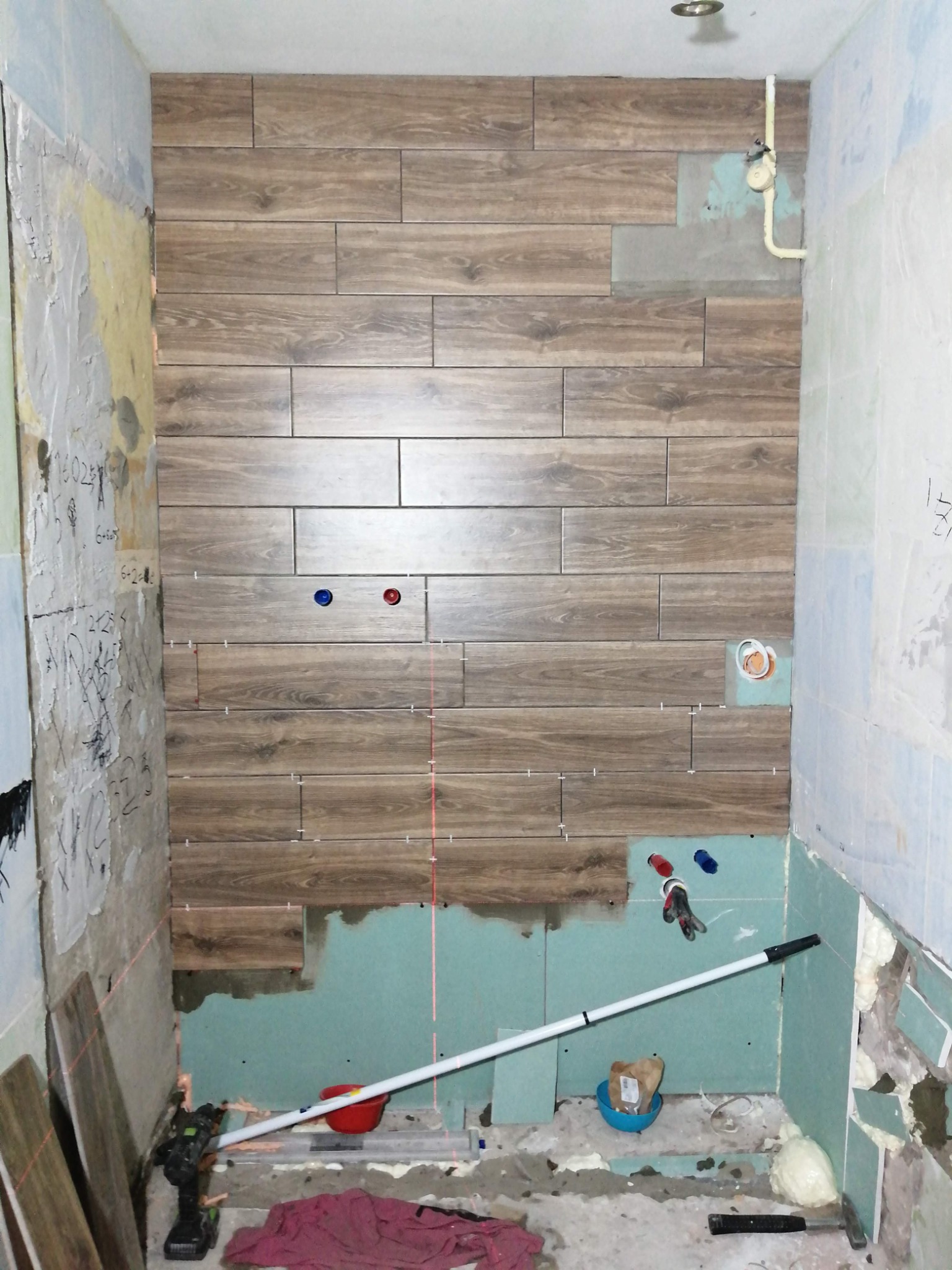Montaż płytek imitujących drewno na ścianie w łazience w trakcie remontu, widoczne narzędzia, poziomica laserowa, nieukończone ściany i instalacje hydrauliczne.