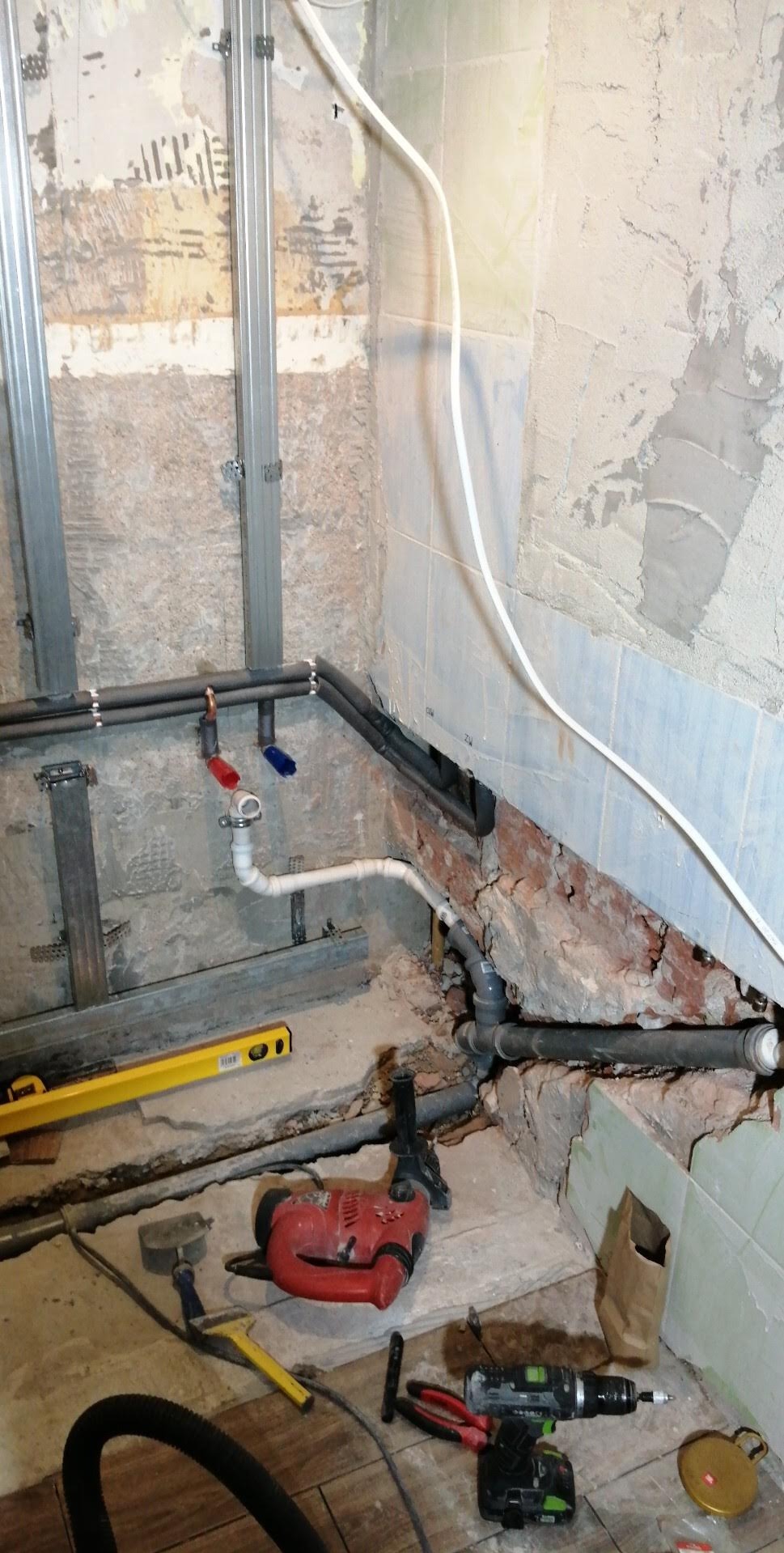 Łazienka w trakcie remontu: widoczne rury instalacji wodnej z zaworami ciepłej i zimnej wody, metalowe profile, ślady po starych płytkach, narzędzia budowlane, w tym wiertarka, poziomica i młotek.