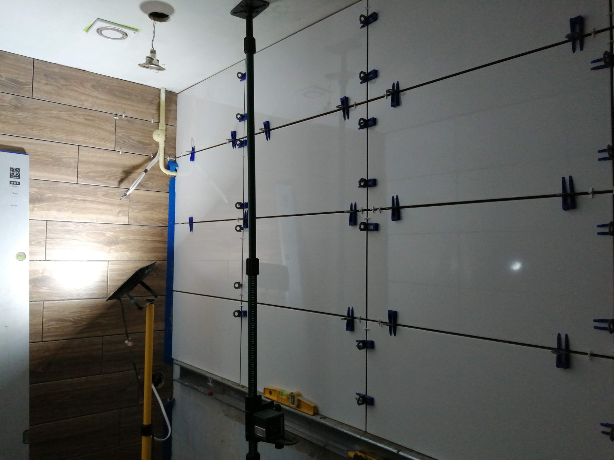 Układanie białych płytek na ścianie z użyciem niebieskich klipsów poziomujących, widoczne narzędzia glazurnicze, fragment ściany z brązowymi płytkami imitującymi drewno.