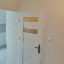 Mark Bud - Białe, przeszklone drzwi wewnętrzne z poziomymi szybami, zamontowane w jasnym pomieszczeniu. Widoczny fragment ściany z włącznikiem światła i fragment podłogi.