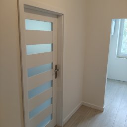 Mark Bud - Białe drzwi wewnętrzne z przeszkleniami w nowocześnie wykończonym wnętrzu. Widoczna podłoga z paneli i fragment okna w sąsiednim pomieszczeniu. Minimalistyczny design.