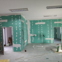 Mark Bud - Wnętrze w trakcie remontu: zielone płyty gipsowo-kartonowe na ścianach, częściowo wyszpachlowane, widoczne kable, drabina, materiały budowlane na podłodze.