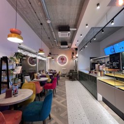 Mark Bud - Wnętrze kawiarni z betonowym sufitem, kolorowymi krzesłami i wzorzystą podłogą. Widoczne lampy wiszące i lada z ekspozycją.