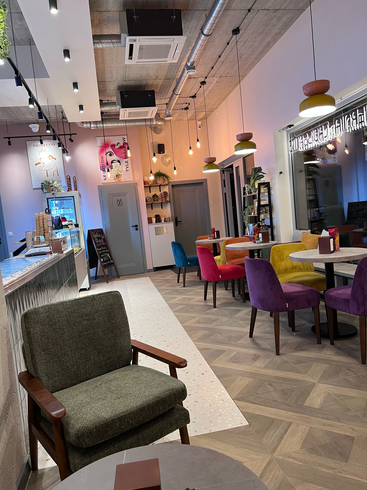 Wnętrze kawiarni z kolorowymi krzesłami i drewnianą podłogą w geometryczny wzór. Widoczne oświetlenie punktowe i dekoracyjne plakaty na ścianach.