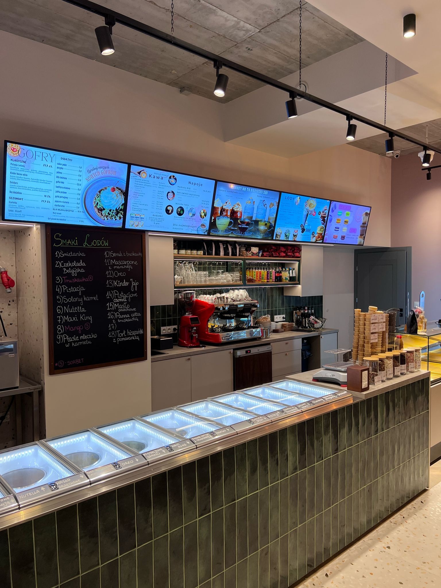 Wnętrze lodziarni z ladą chłodniczą, tablicą smaków lodów i menu na ekranach. Widoczne ekspresy do kawy i zapasy wafelków. Nowoczesny design.