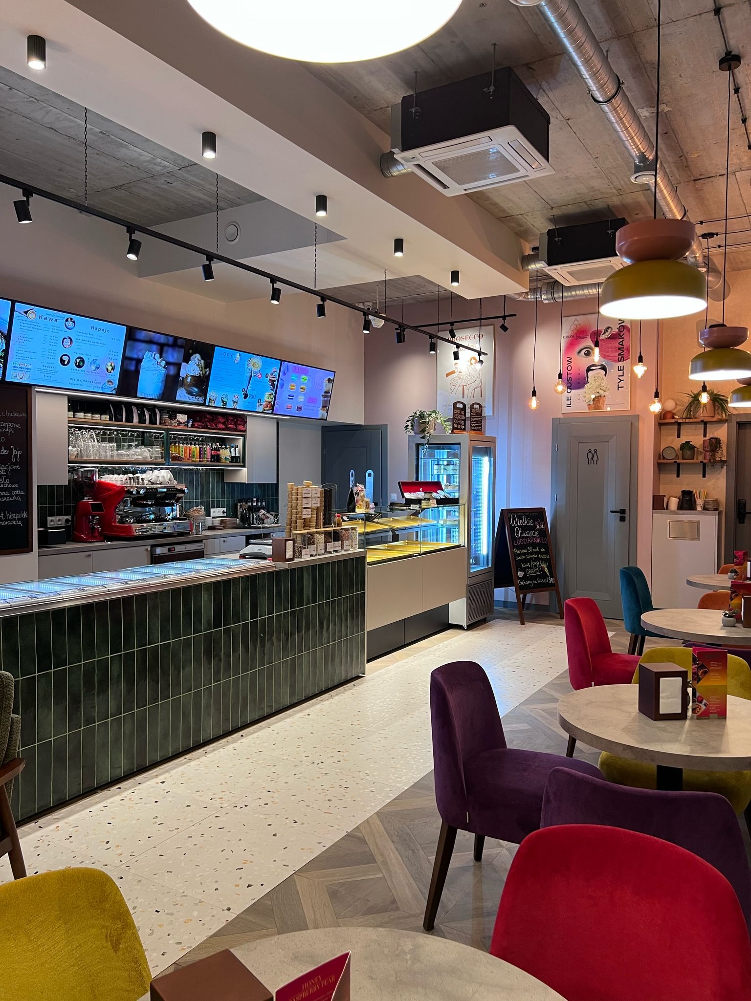 Wnętrze kawiarni z zieloną ladą, ekspresami do kawy i stolikami z kolorowymi krzesłami. Widoczne menu na ekranach i tablica z ofertą lodów.