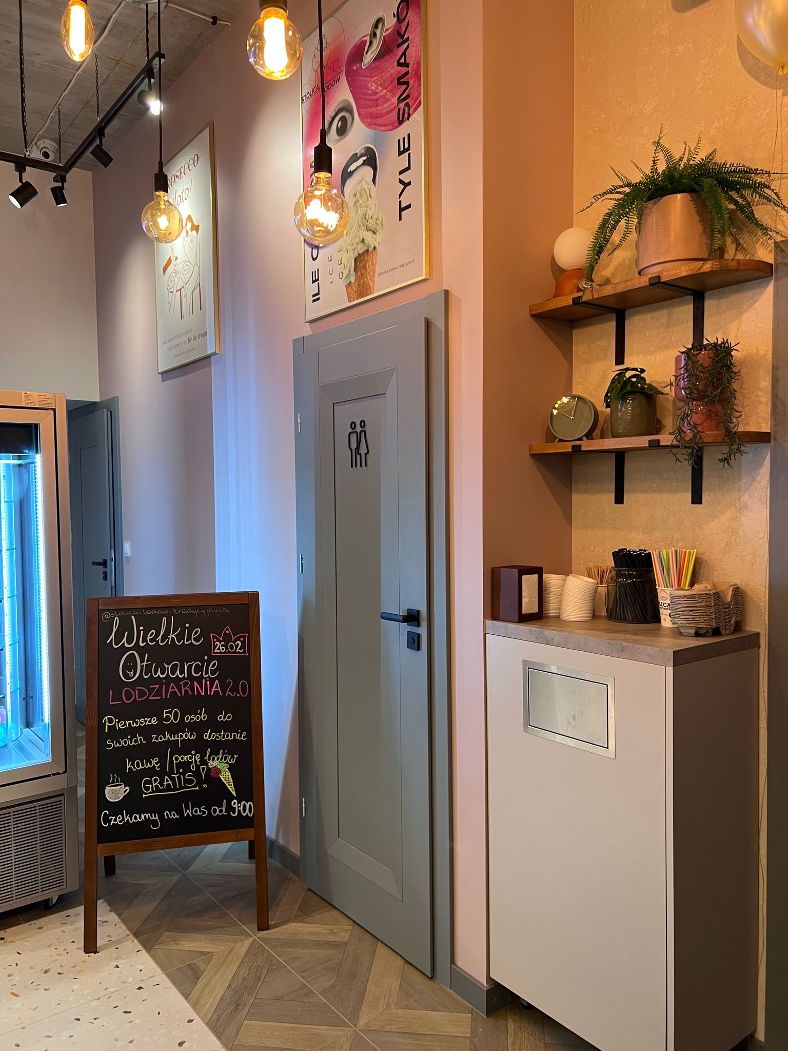Wnętrze lodziarni: tablica z ogłoszeniem otwarcia, drzwi do toalety z oznaczeniem płci, półki z roślinami i akcesoriami do lodów. Ciepłe oświetlenie, pastelowe kolory ścian.