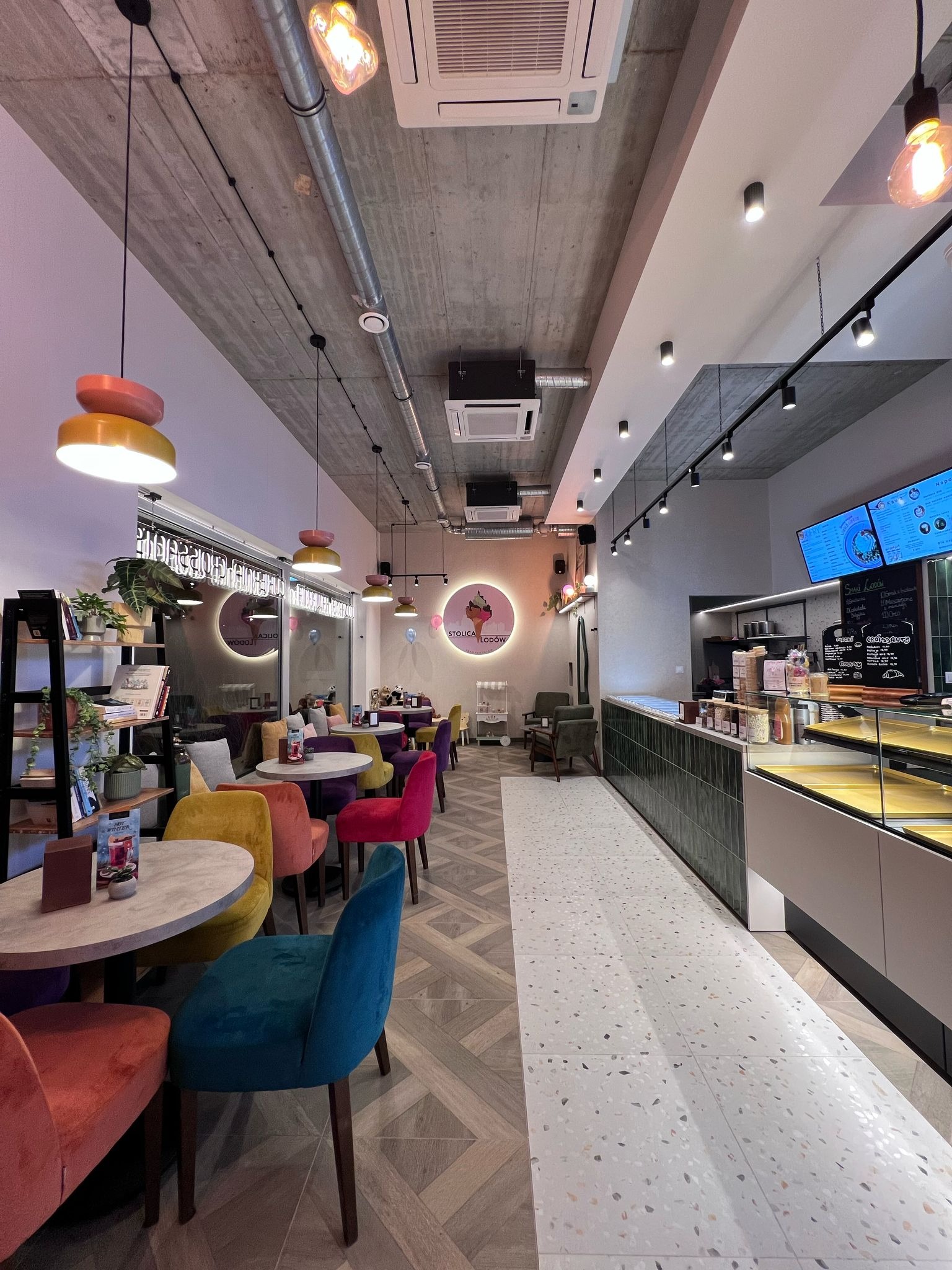 Wnętrze kawiarni z betonowym sufitem, kolorowymi krzesłami i wzorzystą podłogą. Widoczne lampy wiszące i lada z ekspozycją.