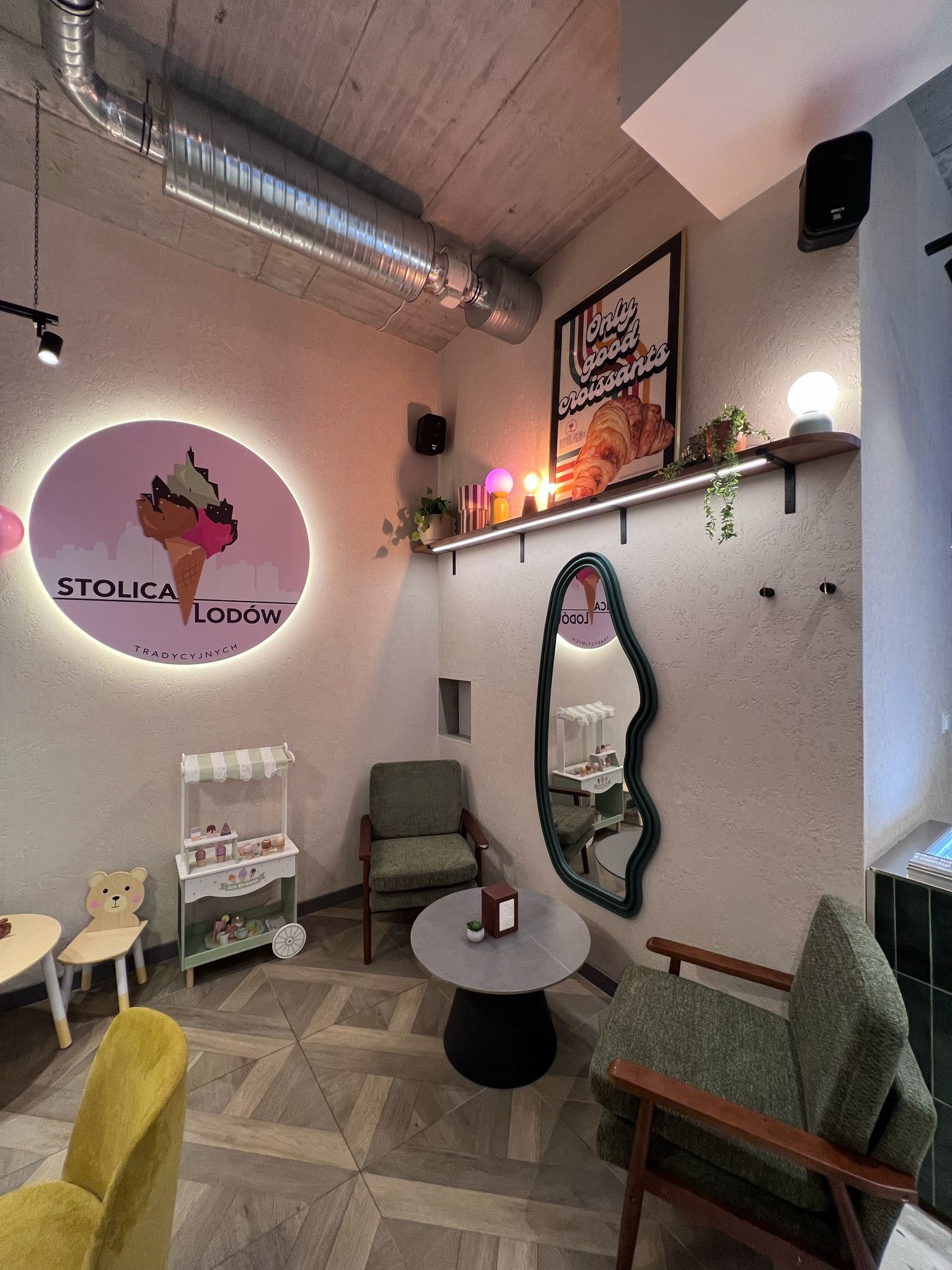 Wnętrze kawiarni z neonem 'Stolica Lodów', stolikami i krzesłami w stylu retro. Widoczne elementy dekoracyjne, takie jak lustro o nieregularnym kształcie i drewniana podłoga.