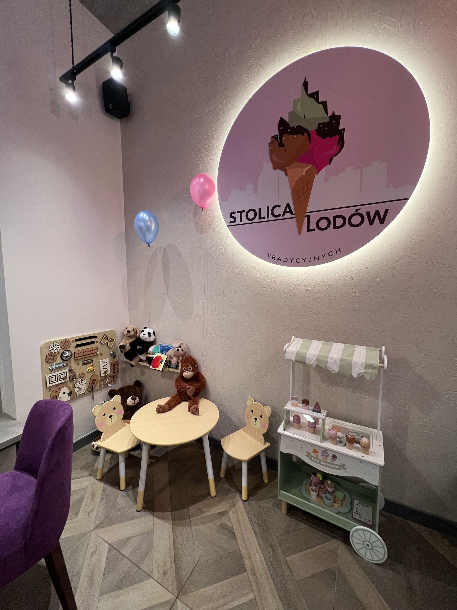 Kącik zabaw dla dzieci w kawiarni: stolik z krzesełkami w kształcie misiów, tablica manipulacyjna, zabawki i podświetlany szyld 'Stolica Lodów'.