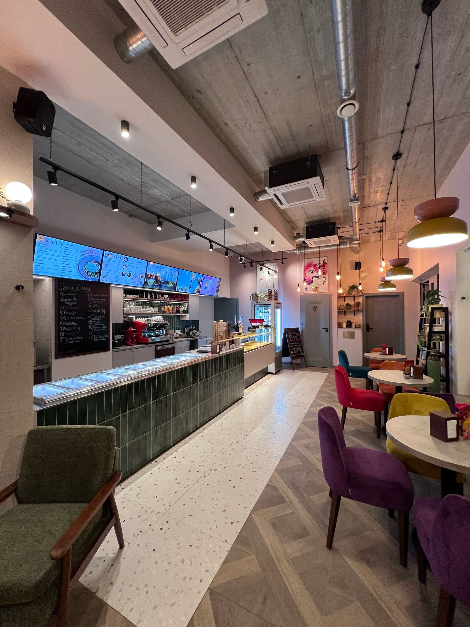 Wnętrze kawiarni z ladą z zielonych płytek, stolikami z kolorowymi krzesłami i menu na tablicy. Widoczne oświetlenie sufitowe i dekoracyjne lampy.