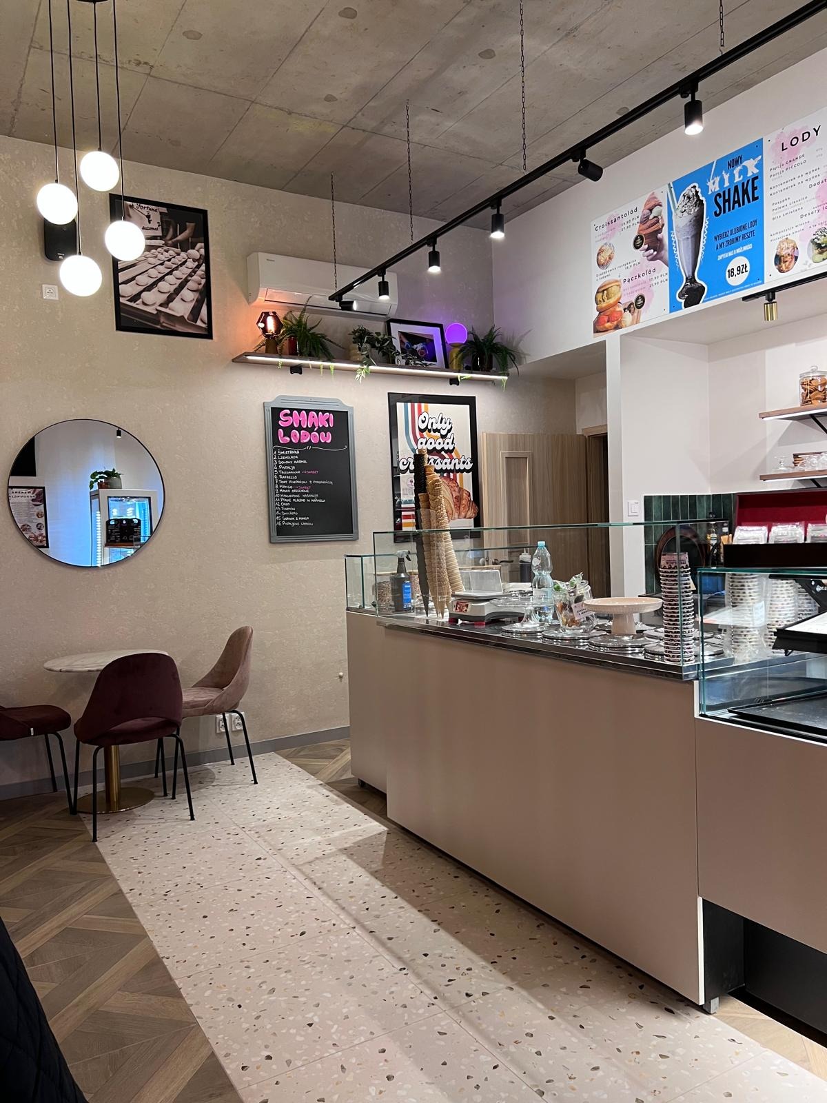 Jasne wnętrze lodziarni z ladą chłodniczą, stolikami i krzesłami. Na ścianach menu i dekoracje. Widoczne oświetlenie punktowe i terazzo na podłodze.