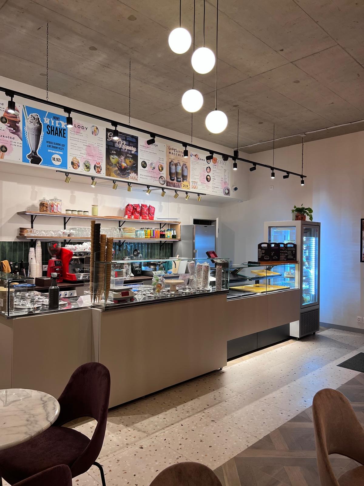 Jasne wnętrze kawiarni z ladą, ekspresami i menu na ścianie. Widoczne stoliki z krzesłami i oświetlenie sufitowe. Nowoczesny design z elementami betonu.