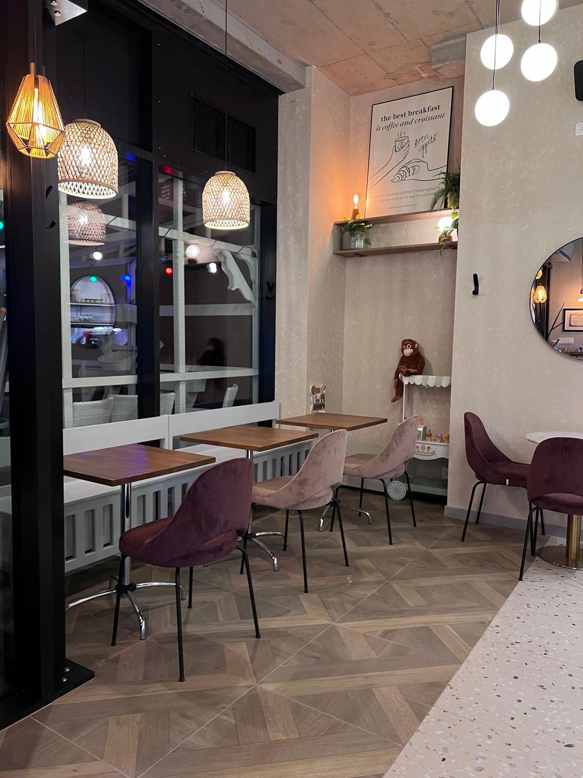 Stylowe wnętrze kawiarni z drewnianą podłogą w jodełkę, stolikami i krzesłami w odcieniach fioletu i beżu. Dekoracje: lampy, plakat i pluszowa małpka.