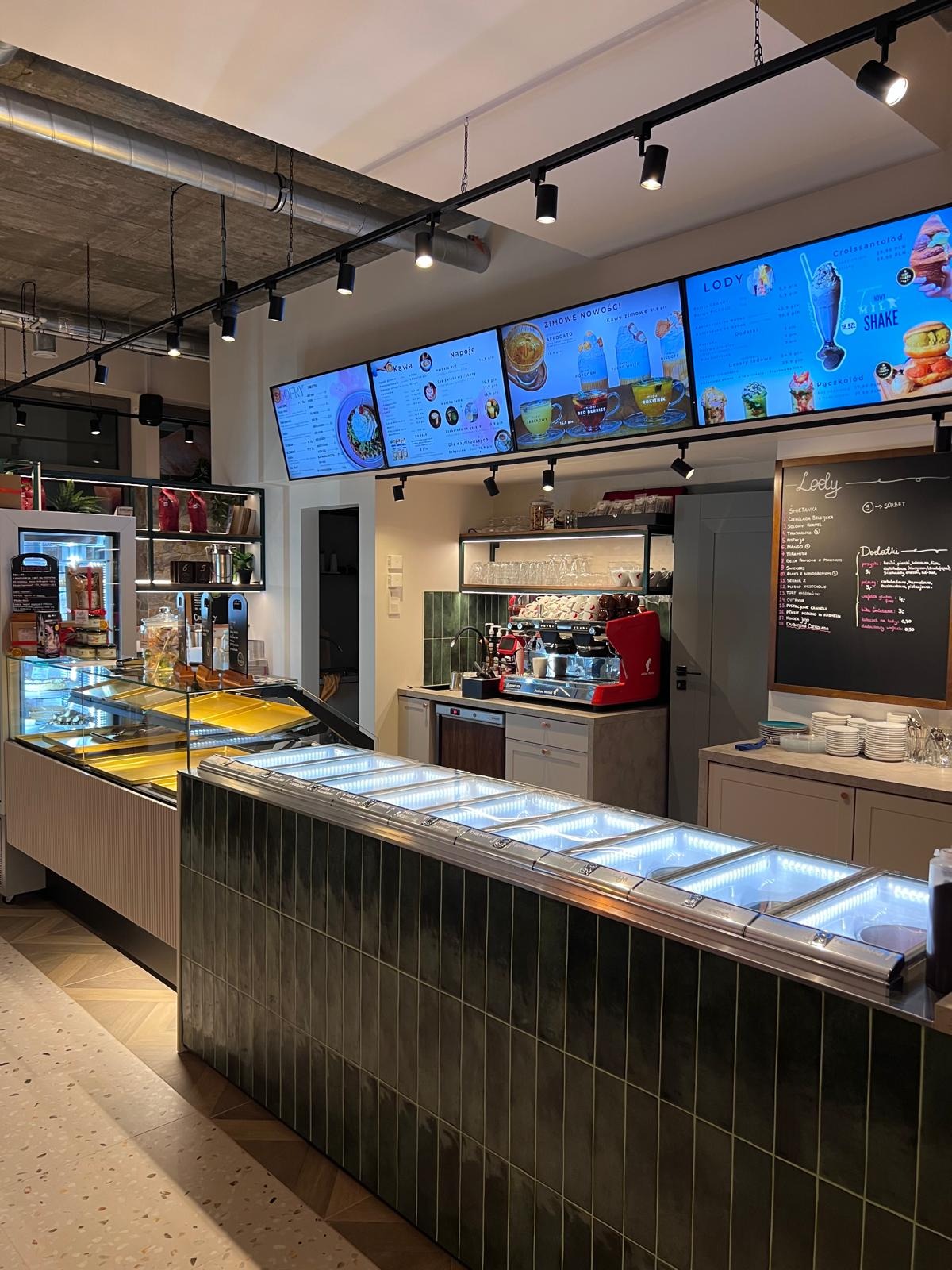 Wnętrze kawiarni z ladą chłodniczą na lody, menu na ekranach i tablicy kredowej. Widoczne ekspresy do kawy i elementy wystroju w stylu minimalistycznym.