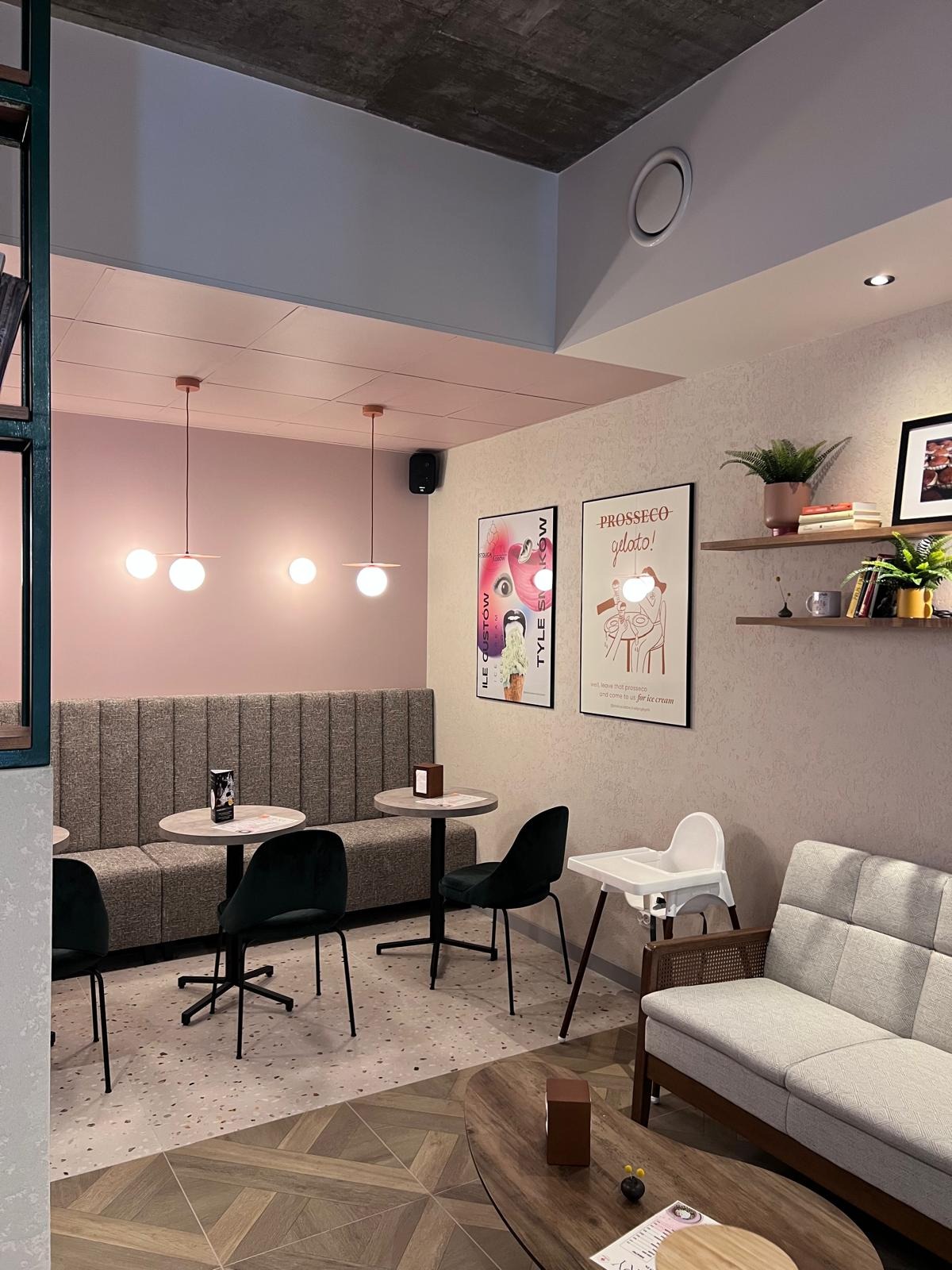 Pastelowe wnętrze kawiarni z tapicerowanymi siedziskami, okrągłymi stolikami i plakatami. Na ścianach półki z roślinami i dekoracjami. Nowoczesny design z elementami retro.